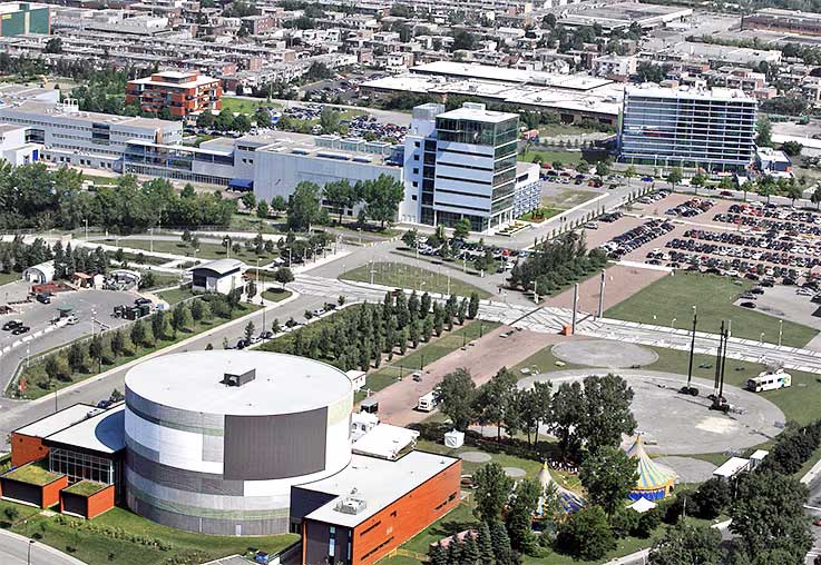 Aerial view of a modern urban area with several multi-story buildings, parking lots, and green spaces. The foreground features a circular building with a flat roof and adjacent rectangular structures. Trees line the streets and pathways throughout the area.