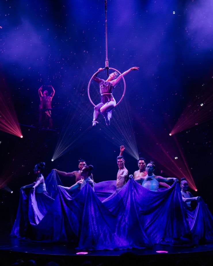 Woman sitting on a suspended hoop with multiple dancers performing underneath. 