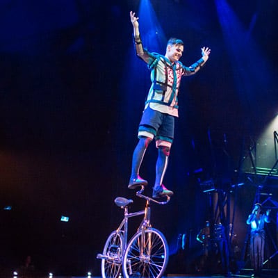 Un artiste en équilibre sur le guidon d'un vélo lève les mains dans les airs - Bazzar Cirque du Soleil