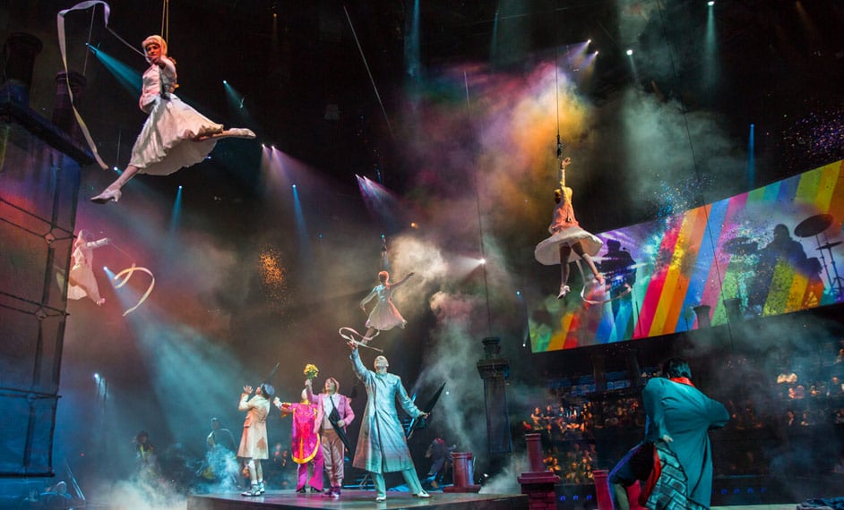 Coloridos personajes miran y señalan a mujeres vestidas de blanco que cuelgan de cuerdas en el aire - The Beatles Love Cirque du Soleil