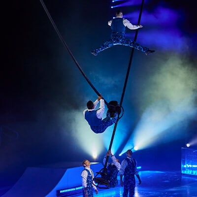 Dos acróbatas se equilibran y saltan sobre mástiles pendulares en un entorno iluminado en azul - Crystal Cirque du Soleil