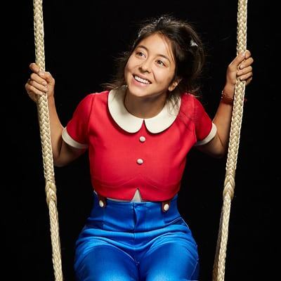 Julie is dressed in a red shirt and blue pants and is sitting on a swing - Cirque du Soleil Orlando