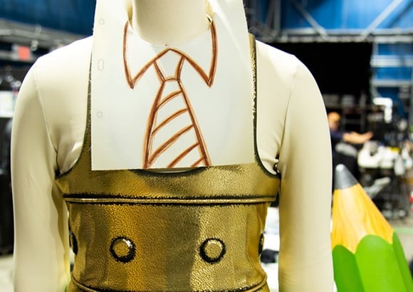 A pencil looking mannequin is wearing a drawn tie on his neck, a golden top and green pants Cirque du Soleil Disney Springs
