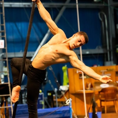 Performer holds to a black pole with one hand while opening his other arm during aerial training - Disney Cirque du Soleil