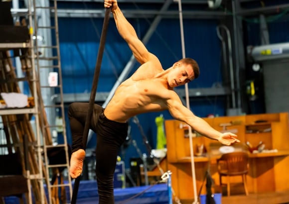 Performer holds to a black pole with one hand while opening his other arm during aerial training - Disney Cirque du Soleil