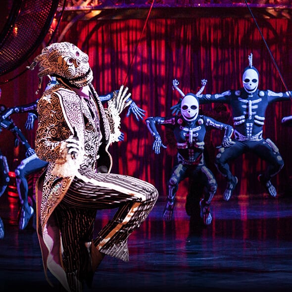 A tribe of skeletons dance together in front of a red curtain - Cirque du Soleil Kooza