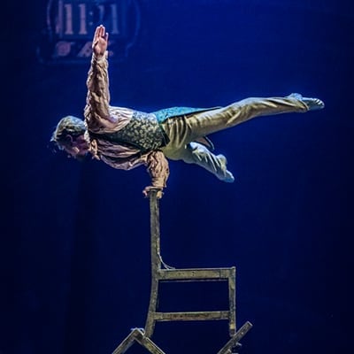 Performer achieves a one hand handstand on an Indian pole that has the appearance of a chair - Kurios cirque