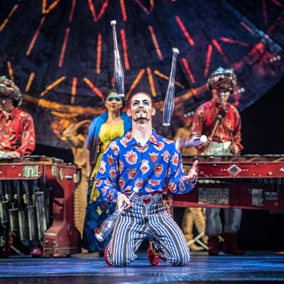 Circus artist juggles with bowling pins while musicians play in the background - cirque Luzia