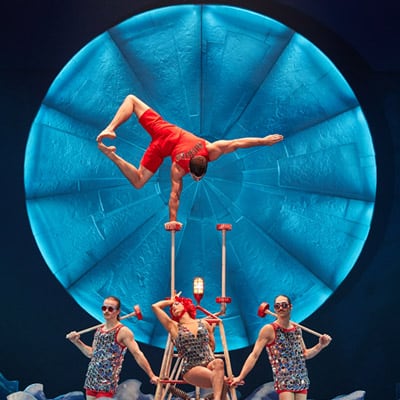 Un artista vestido de rojo se equilibra con una mano en frente de un gran disco - Luzia Cirque du Soleil