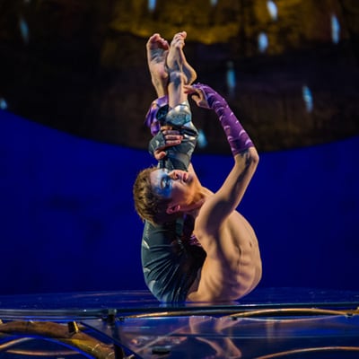 El contorsionista sostiene sus piernas detrás de su espalda sobre una mesa de vidrio - Luzia Cirque du Soleil
