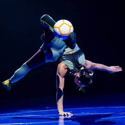 Un artista vestido con ropa deportiva hace una parada de manos en una mano con una pelota de fútbol entre los pies - Messi10 Cirque du Soleil