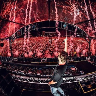 Un DJ toca música frente a una inmensa multitud iluminada en rojo mientras el humo comienza a cubrir el escenario - MŪV Circo del Sol