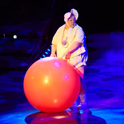 Un payaso vestido con ropa de bebé sostiene una bola naranja de gran tamaño frente a él - Mystère Cirque du Soleil
