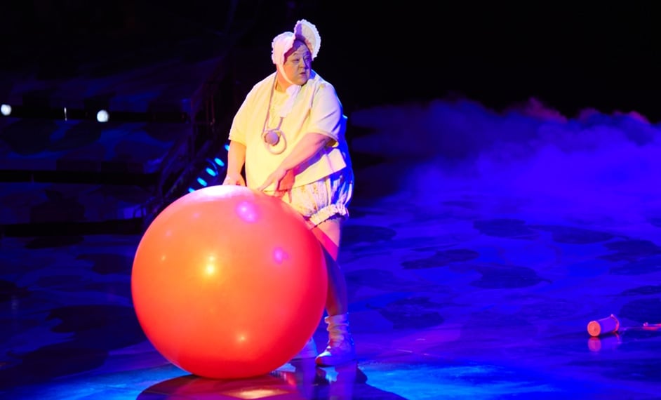 Un payaso vestido con ropa de bebé sostiene una bola naranja de gran tamaño frente a él - Mystère Cirque du Soleil