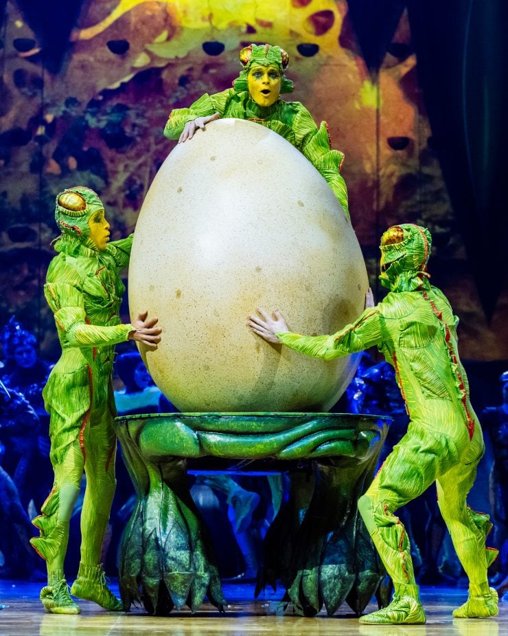 Tres acróbatas vestidos con trajes de insectos verdes sostienen un huevo gigante. 