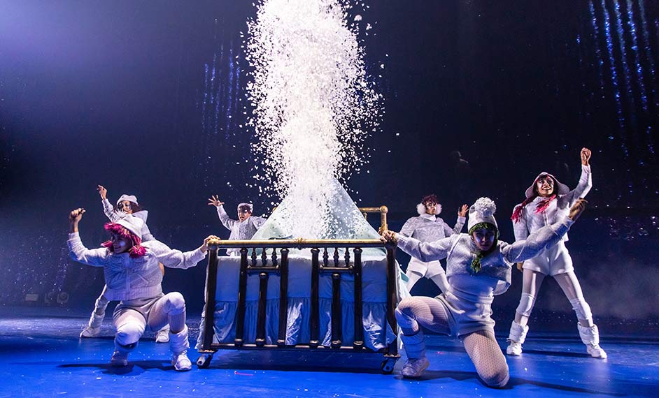 Six mischievous elves dressed in white clothing dance around a bed - Cirque du Soleil Twas The Night Before show